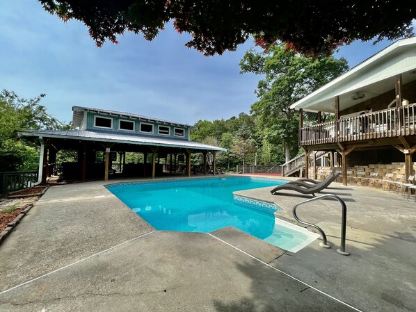 A swimming pool with a covered deck area, a wooden deck with chairs, and a building with a balcony.