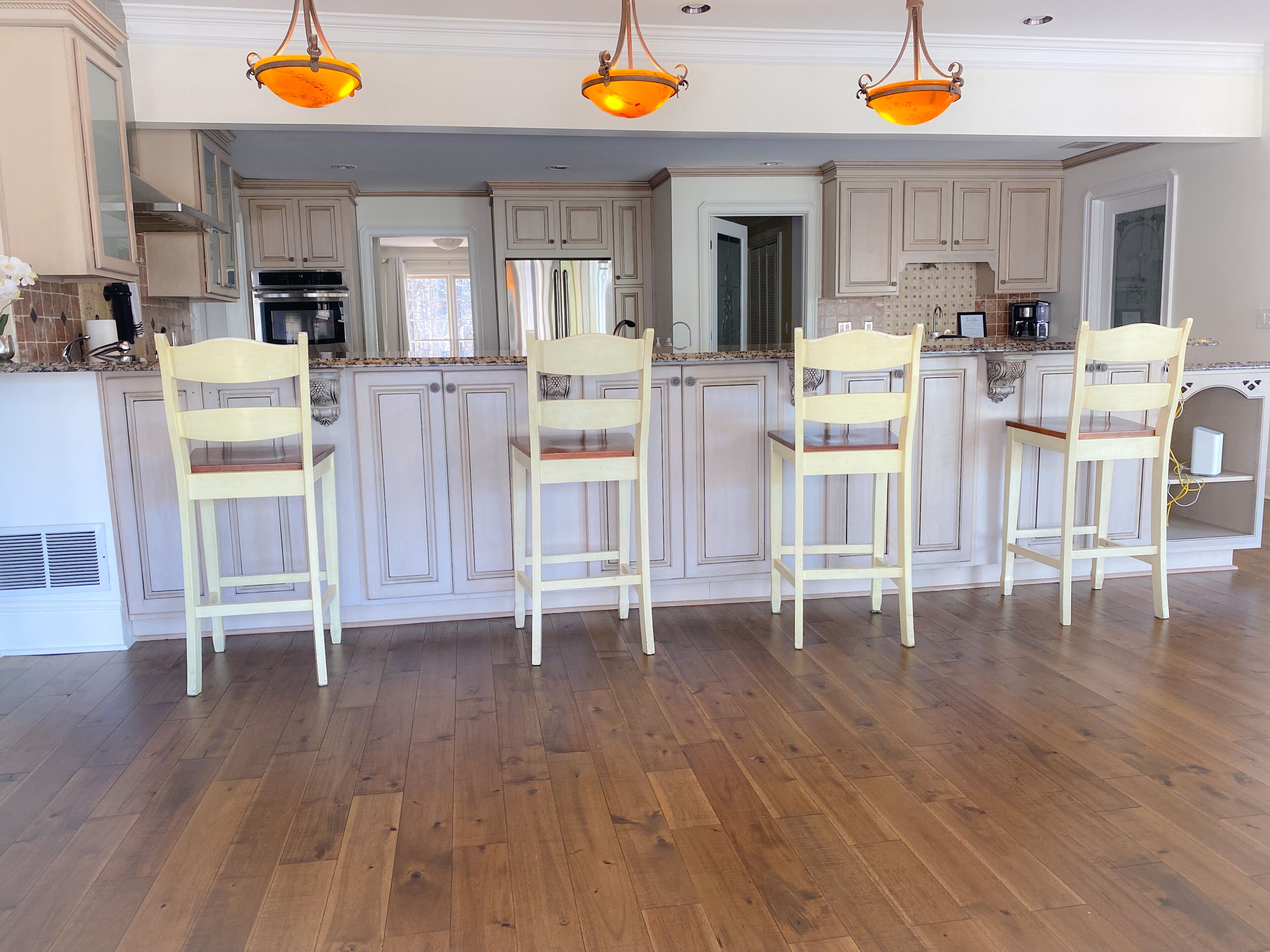 Cocina con suelo de madera, armarios blancos y tres taburetes de bar.