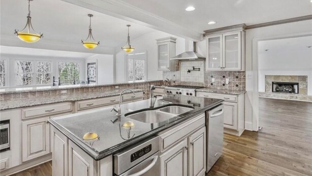 Una cocina amplia con armarios blancos, una isla grande con un lavamanos y un horno empotrado. Hay luces colgantes sobre la isla y una chimenea al fondo.