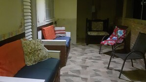 A room with a brick wall, a window with blinds, a sofa with cushions, a chair, and a patterned floor. - Refúgio da chapada (Cavalcante)