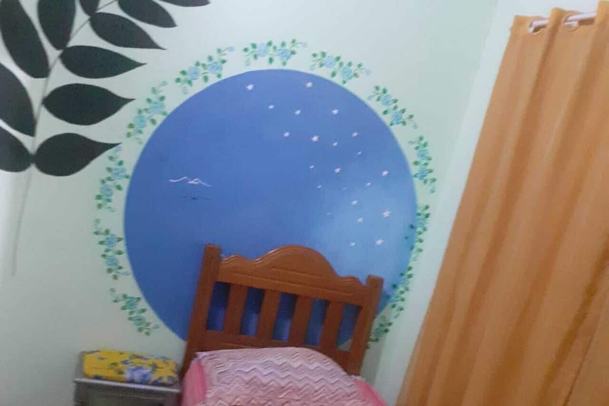 A small bedroom with a bed, a wooden headboard, a blue wall decoration, a curtain, and a small table with items on it.