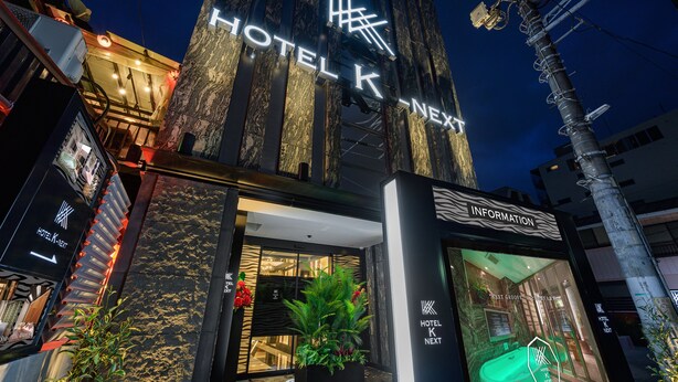A hotel entrance with a modern design, a visible reception area, and a sign indicating the hotel name. - HOTEL K-NEXT (Saitama)
