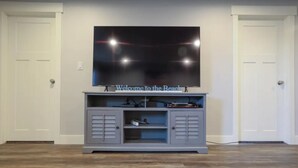 A living room with a flat-screen TV mounted on a grey entertainment unit, a door on the left, and a door on the right. - Holiday Haven | 7BR Sleeps 24 | Private Pickleball Court, Lake Views & Beach Access (Garden City)