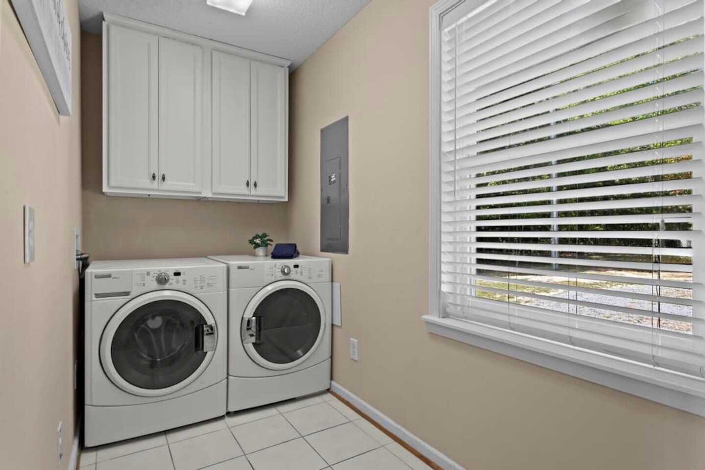 A laundry room with two front-loading washing machines, a cabinet above, and a window with blinds.