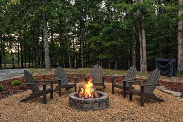 Un foyer circulaire en pierre, avec un feu allumé, entouré de quatre chaises Adirondack.