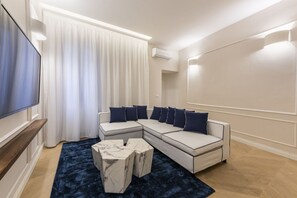 A modern living room with a sectional sofa, a flat-screen TV, a marble coffee table, and a dark blue rug.