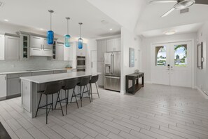 Una cocina moderna con armarios blancos, una isla central con taburetes de bar, electrodomésticos de acero inoxidable y lámparas colgantes azules.