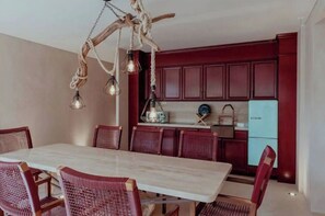 Un comedor con mesa de mármol y sillas de mimbre, una lámpara rústica y una cocina con armarios rojos y un refrigerador antiguo.