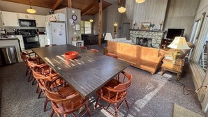 A spacious kitchen with a large wooden table and chairs, a stone fireplace, and a dining area with a sofa and lamp.