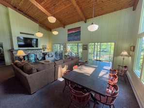 A spacious living area with a large sofa, a dining table, and multiple chairs. There is a fireplace with a stone surround, a television mounted on the wall, and a view of the outdoors through large windows.