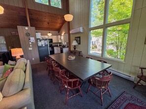 A dining area with a long table and multiple chairs, a kitchen in the background, and large windows offering a view of greenery.