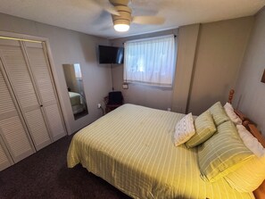 Un dormitorio con cama, ventilador de techo, televisor, ventana con cortinas y armario.