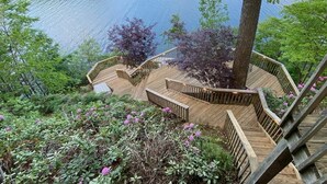A wooden deck with a spiral staircase leading to a platform over a body of water, surrounded by lush greenery and flowering plants.