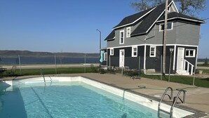 A rectangular swimming pool with clear blue water, surrounded by a concrete deck. There are metal pool ladders on the deck. In the background, there is a two-story house with a dark gray exterior and white trim. The house has a small porch with outdoor furniture. A body of water is visible in the distance. - Sunset Motel (Lake City)