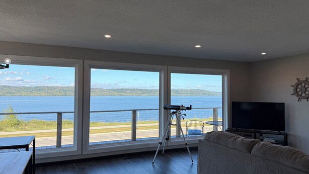 A room with large windows overlooking a lake, a telescope on a tripod, a television, and a sail wheel decoration. - Sunset Motel (Lake City)