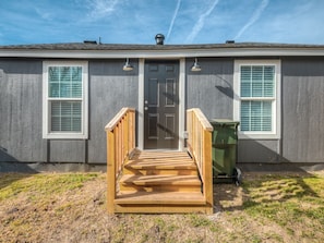 A house with a front door, two windows with shutters, a wooden front porch, and a green trash bin.