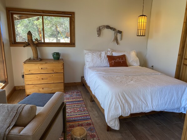 A bedroom with a wooden dresser, a bed with white bedding, a colorful rug, and a window with a view of trees. - Scenic cabin in Carson National Forest (Ranchos de Taos)
