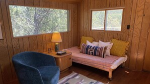 A cozy room with wooden walls, a bed with patterned bedding, a blue chair, and a nightstand with a lamp. - Scenic cabin in Carson National Forest (Ranchos de Taos)