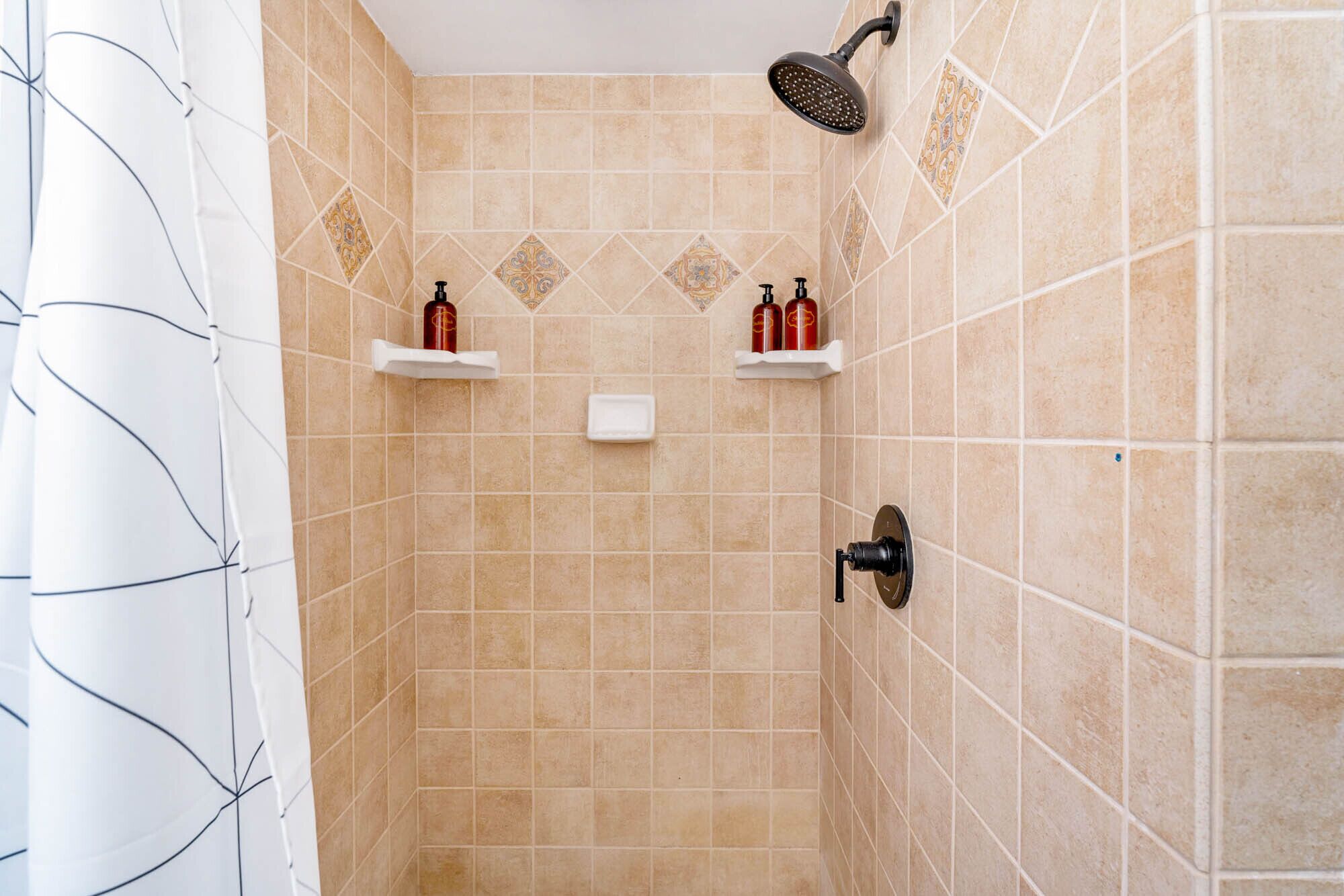 Un espace douche avec des carreaux de couleur beige, une douchette à main et deux étagères blanches sur lesquelles sont posées des bouteilles rouges.