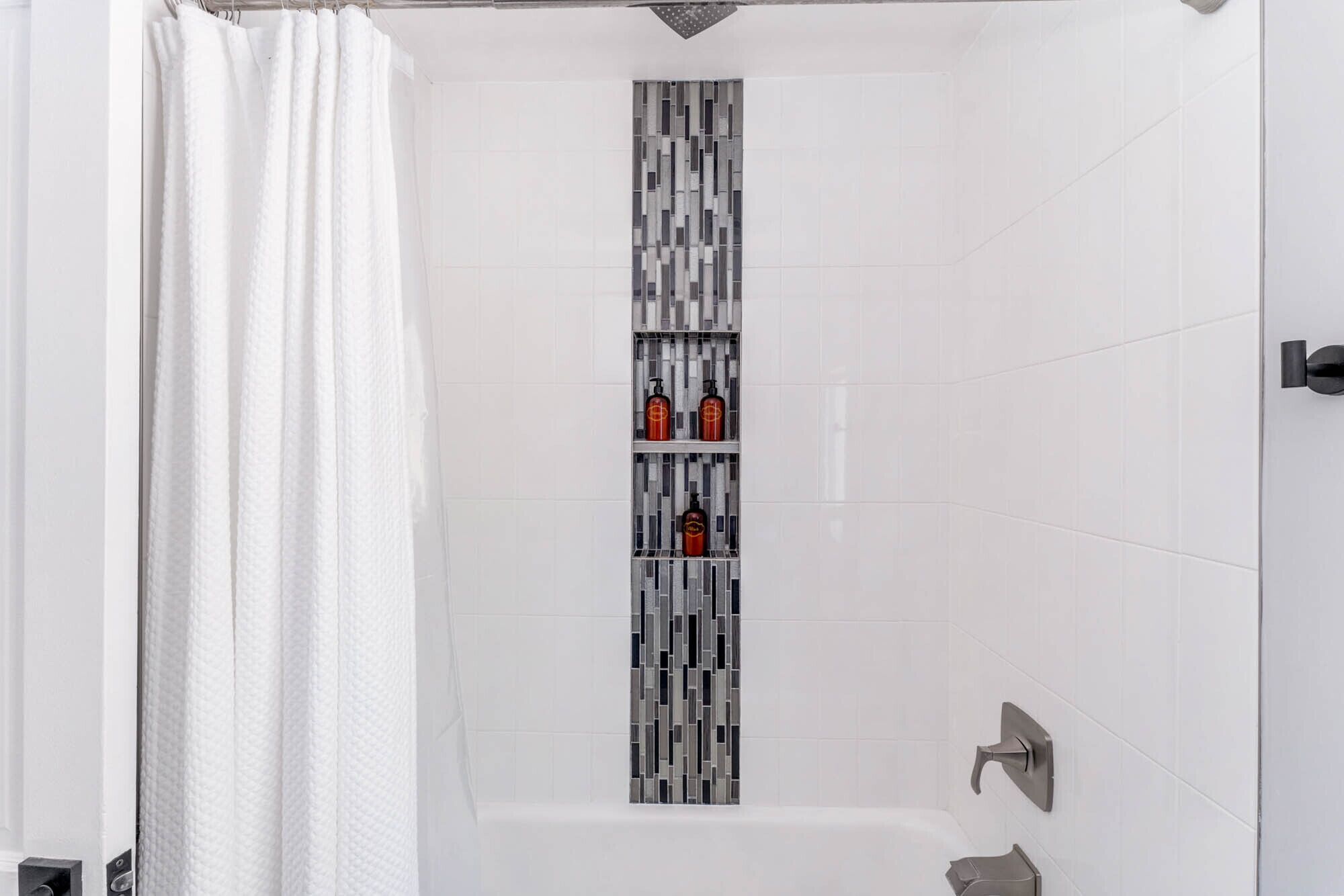 Een douche met een witte douchegordijn, een betegelde wand met een verticaal mozaïekpatroon en een plank met twee flessen.