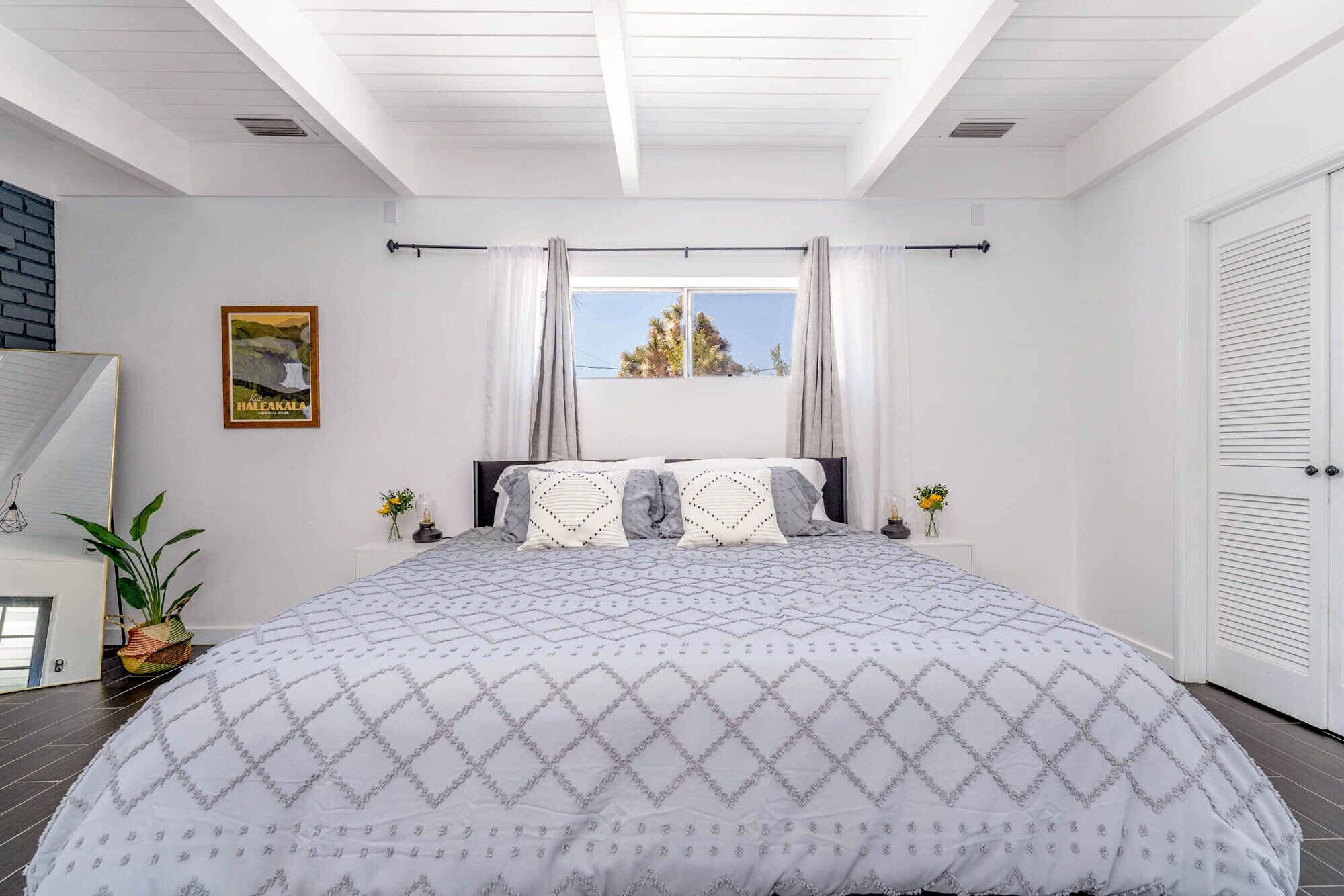 Une chambre avec un grand lit, deux tables de chevet, une fenêtre avec des rideaux, un tableau encadré au mur et une porte à motifs horizontaux.