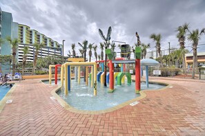 A water play area with colorful structures and a pool, surrounded by palm trees and a brick walkway.