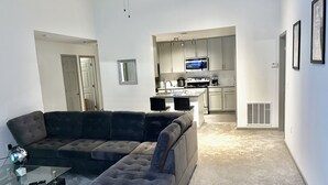 A living room with a sectional sofa, a glass coffee table, and a ceiling fan. A kitchen with white cabinets and appliances is visible in the background.