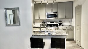 A compact kitchen with a microwave, stove, and coffee maker, featuring two bar stools and a granite countertop.
