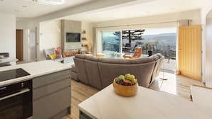 A modern kitchen with a built-in oven, a dining area with a wooden table and a bowl of fruit, and a living room with a sofa and a view of the outdoors. - Vikings Annexe, Minehead (Minehead)