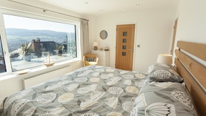 A bedroom with a large window, a bed with patterned bedding, a bedside table with a lamp, and a view of the countryside. - Vikings Annexe, Minehead (Minehead)