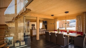 Een ruime woonkamer met een houten trap, een eettafel met stoelen, een keukenhoek en een groot raam met uitzicht op het omliggende landschap.