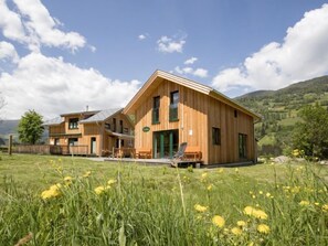 A wooden cabin with a large porch, surrounded by a grassy field and mountains in the background.