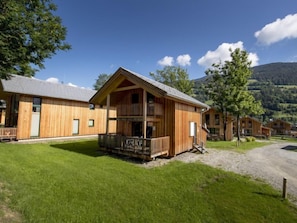 A wooden lodge with a balcony, surrounded by a gravel path and lush greenery, set against a backdrop of mountains.