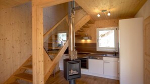 Interior de madera con una escalera, un estufa de leña, una cocina con armarios blancos y un ventanal.