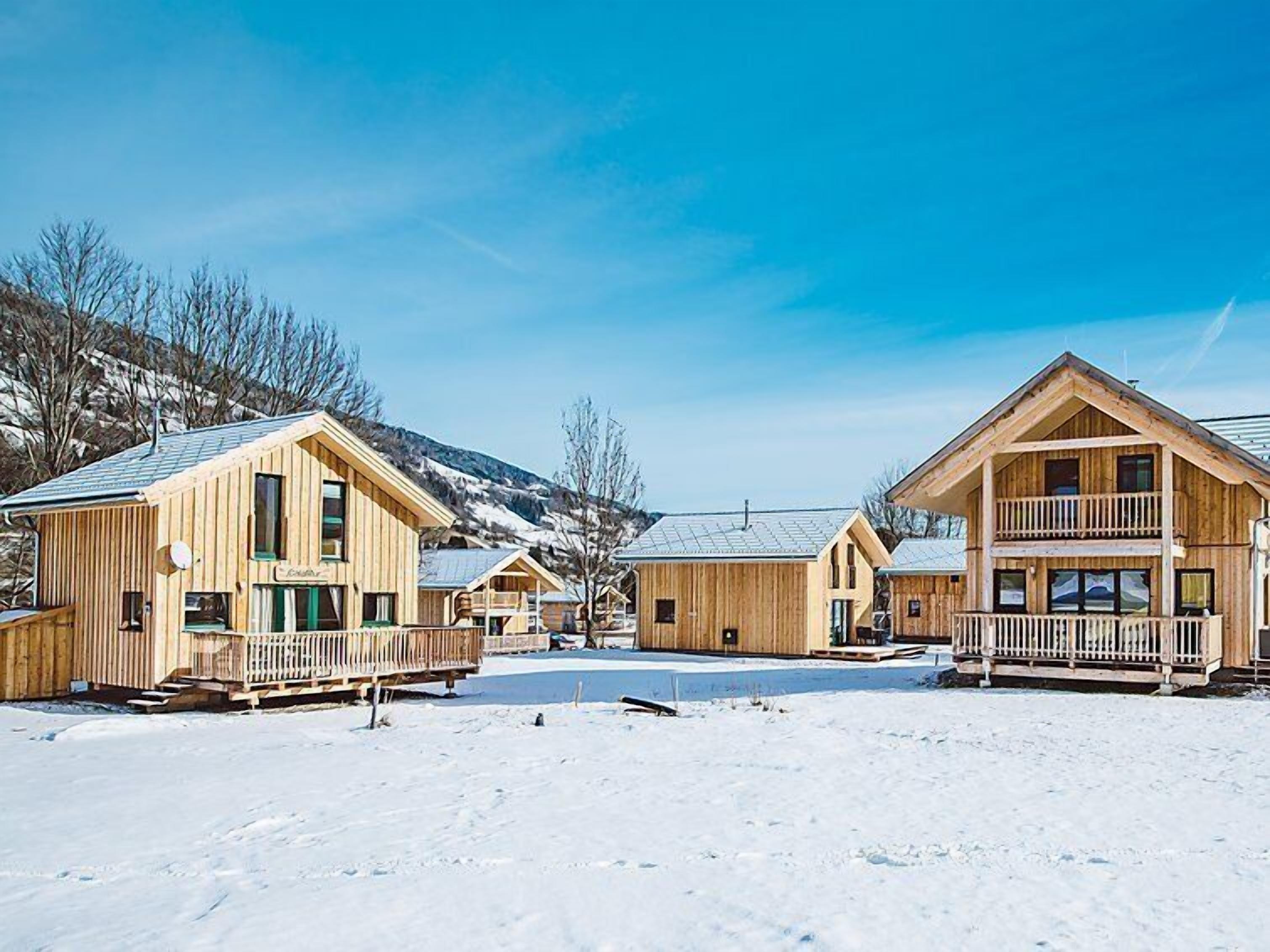 A row of wooden cabins with balconies, set against a snowy landscape with mountains in the background.