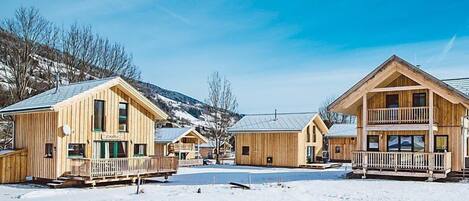 A row of wooden cabins with balconies, set against a snowy landscape with mountains in the background.