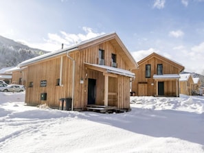 A wooden cabin with a porch, a small attached building, and a snow-covered landscape with mountains in the background.