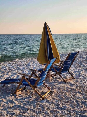 Twee strandstoelen met een gele parasol op een zandstrand, met de oceaan op de achtergrond.