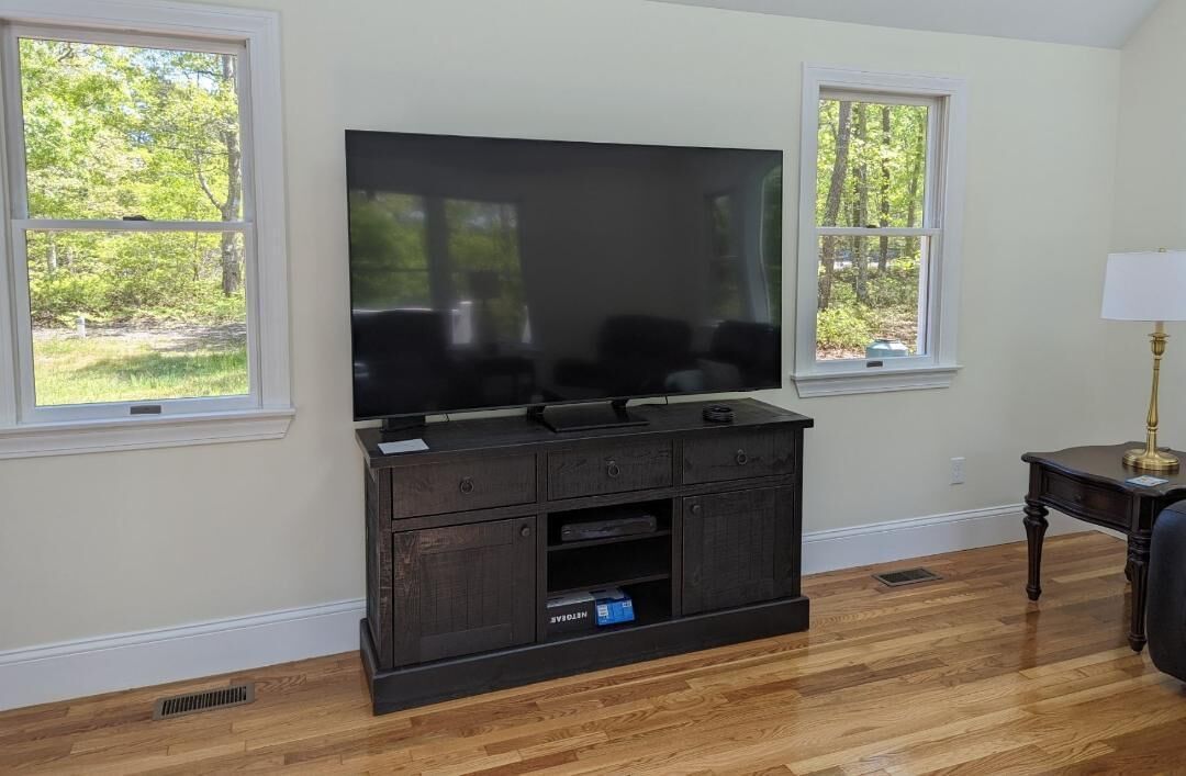 A living room with a large flat-screen TV on a wooden entertainment center, two windows with a view of greenery, and a lamp on a side table.