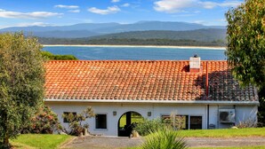 A coastal house with a red-tiled roof, a small garden, and a view of the ocean and mountains. - Cordova - Perfect Summer or Winter Getaway (Merimbula)