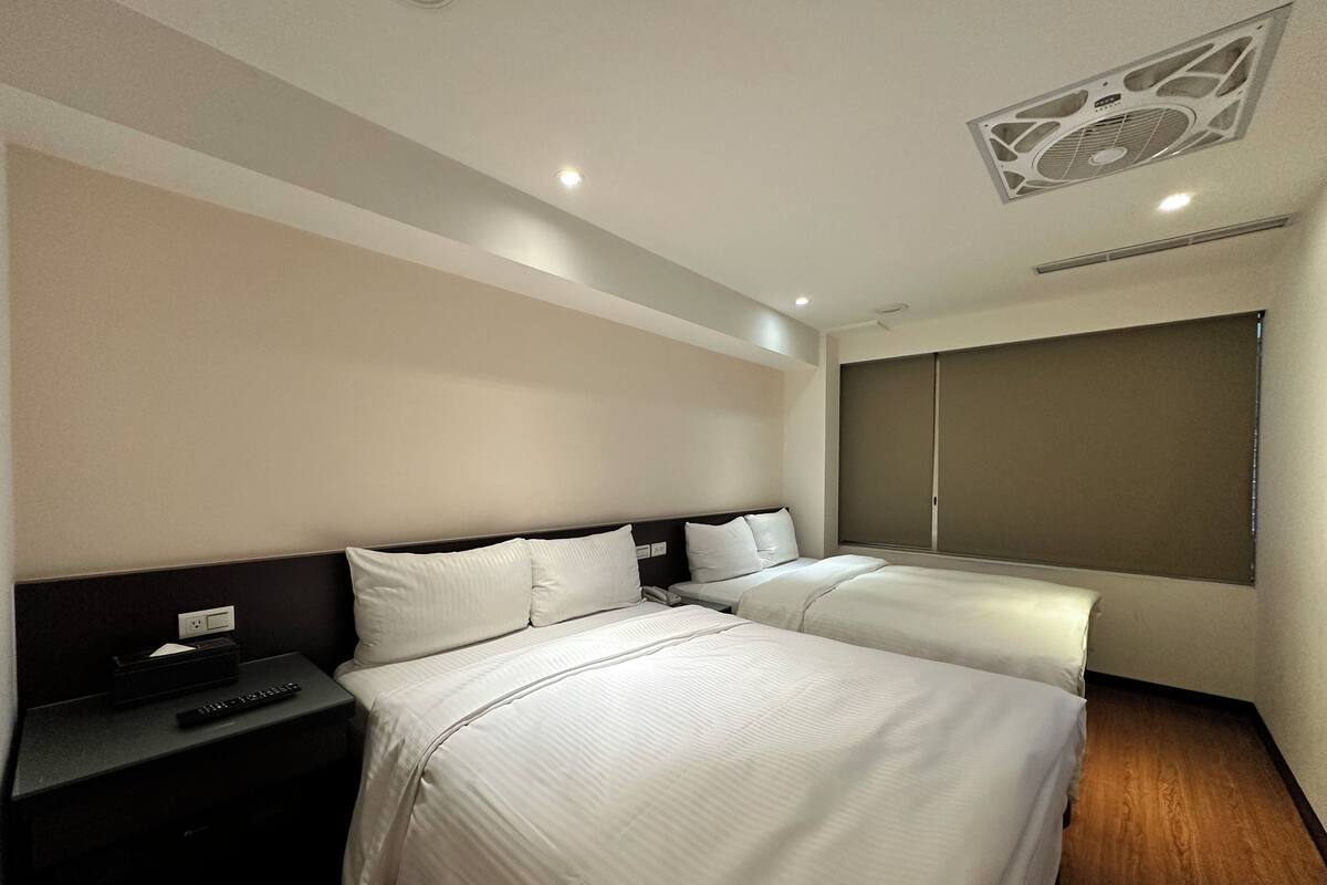Ein Hotelzimmer mit zwei Betten, einem Nachttisch, einem Schreibtisch und einem großen Fenster mit Jalousien.