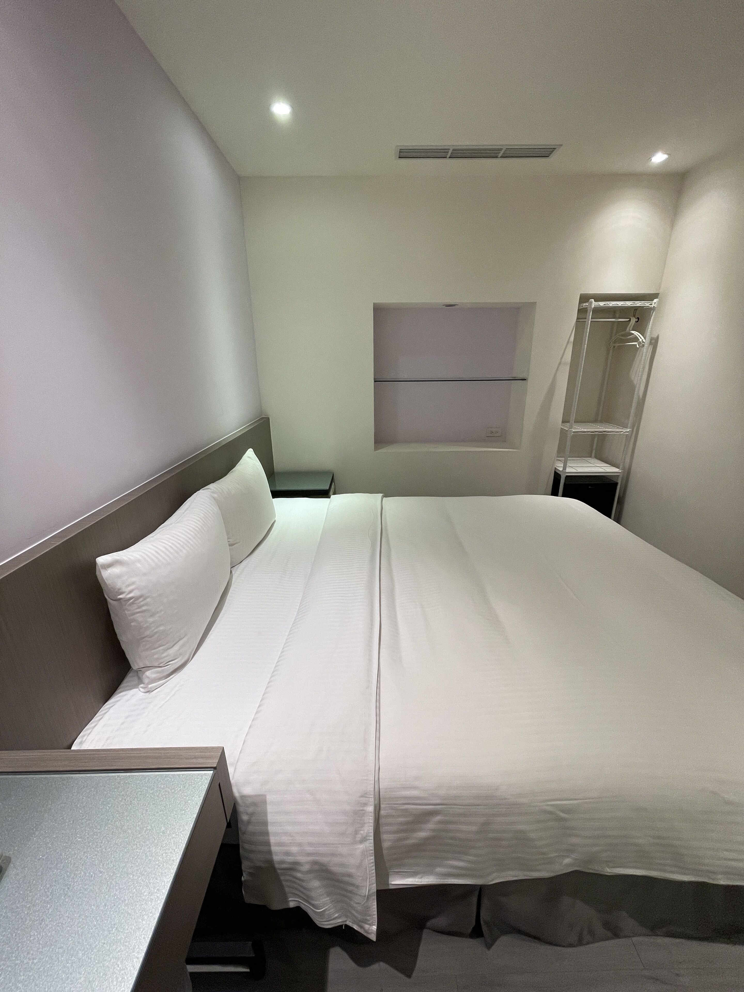 A modern hotel room with a neatly made bed, a small bedside table, a wall-mounted shelf, and a window with a view.