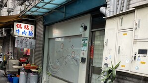 A narrow street with a closed shop featuring a blue awning and a closed shutter door. There are potted plants along the sidewalk and a scooter parked in front. - Good Day Inn (Taipei)