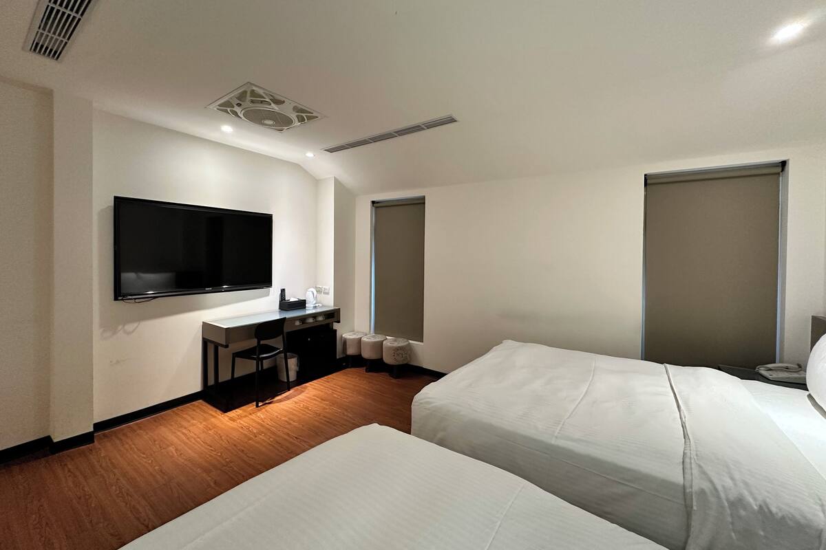 Ein Hotelzimmer mit zwei Betten, einem Schreibtisch mit Stuhl, einem an der Wand befestigten Flachbildfernseher und einem Fenster mit Blick.