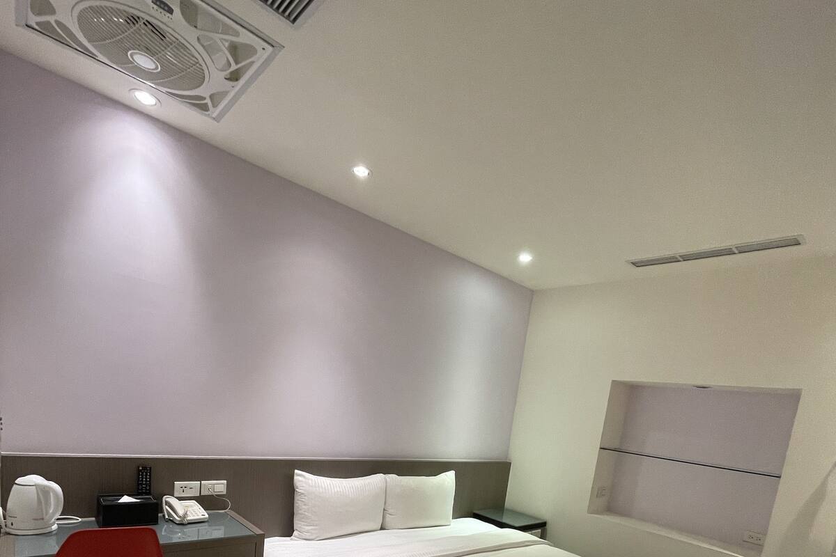 Ein modernes Hotelzimmer mit einem großen Bett, einem Schreibtisch mit Stuhl, einer kleinen Mülleimer und einem Fenster mit Jalousien.