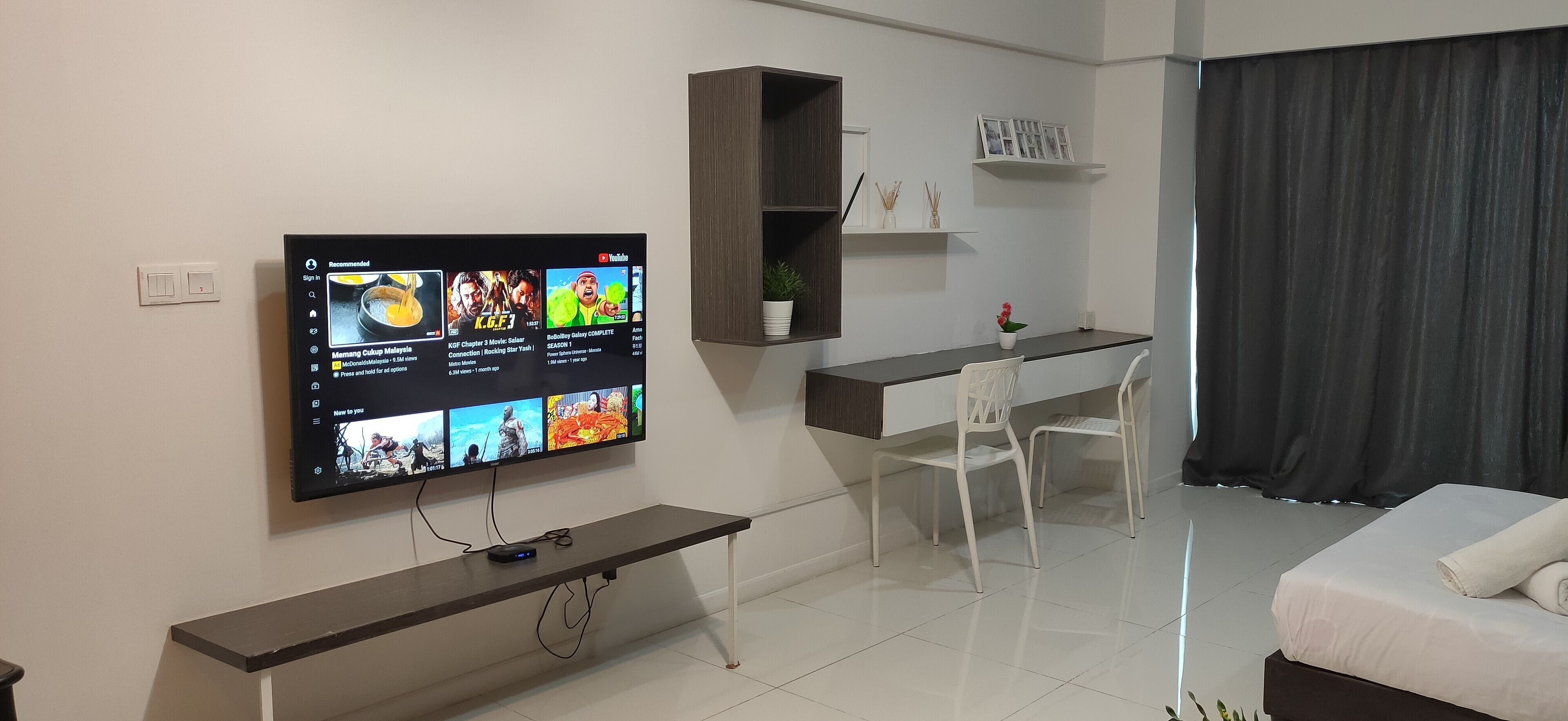 Una moderna camera d'albergo con TV a schermo piatto fissata al muro, una scrivania con una sedia, un letto con un asciugamano arrotolato e una tenda scura.