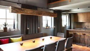 Un espace repas moderne avec une longue table en bois, des chaises à revêtement gris et un coin bar avec des tabourets.