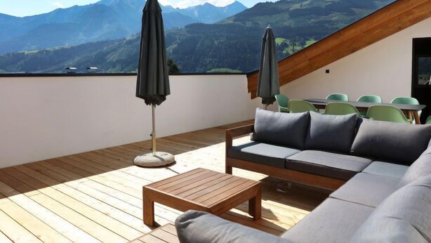 Une terrasse en bois avec un canapé et des fauteuils, une table et des parasols, offrant une vue sur un paysage montagneux.