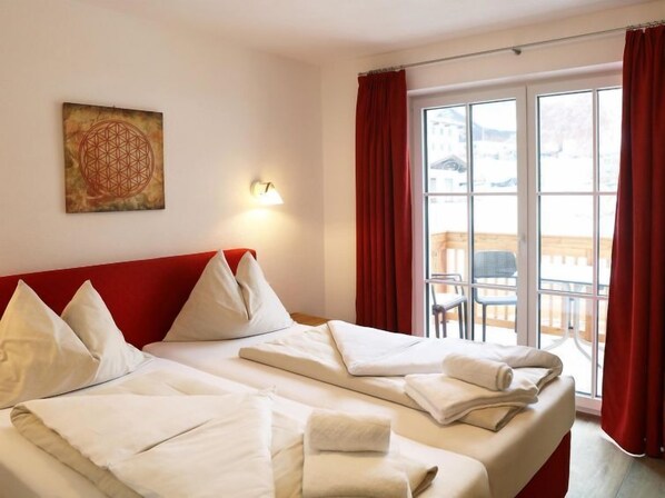 A hotel room with two beds, a red headboard, a wall-mounted light, and a large window with red curtains.