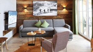 Ein modernes Wohnzimmer mit einem grauen Sofa, zwei grünen Kissen, einem an der Wand befestigten Fernseher und einem großen Bilderrahmen mit einer Berglandschaft.